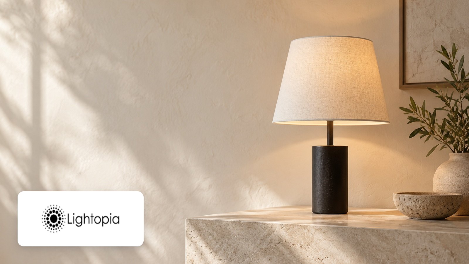 Lightopia logo with Lit table lamp, linen shade, black cylinder base, travertine console, olive sprig